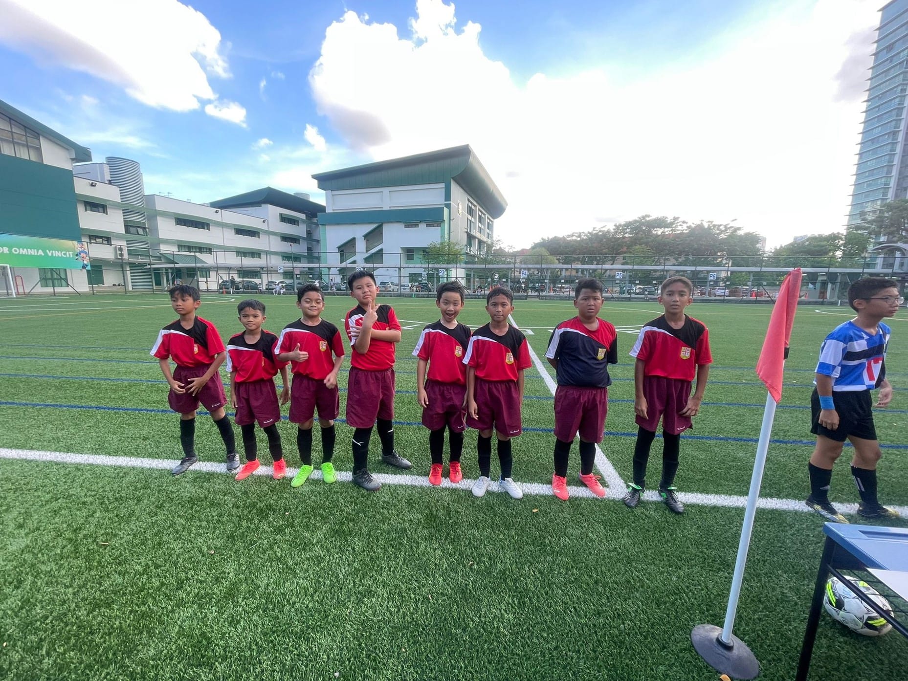Eight young boys in red and black football uniforms line up on a green turf pitch near a red corner flag.
